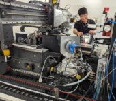 In the foreground, a large metal piece of equipment with many wires and sensors protruding. In the background, a man smiles while placing an object on the instrument.
