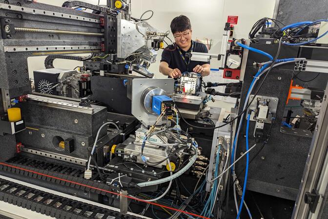 In the foreground, a large metal piece of equipment with many wires and sensors protruding. In the background, a man smiles while placing an object on the instrument.